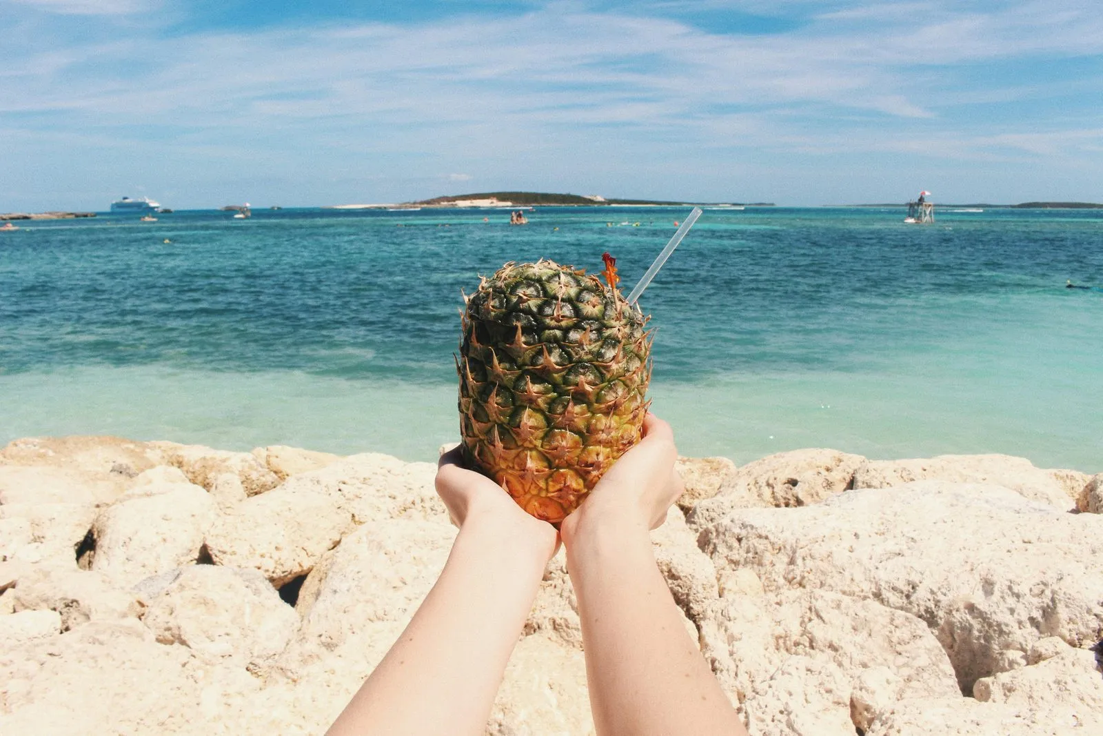 A pineapple drink held up against turquoise water with a cruise ship on the horizon