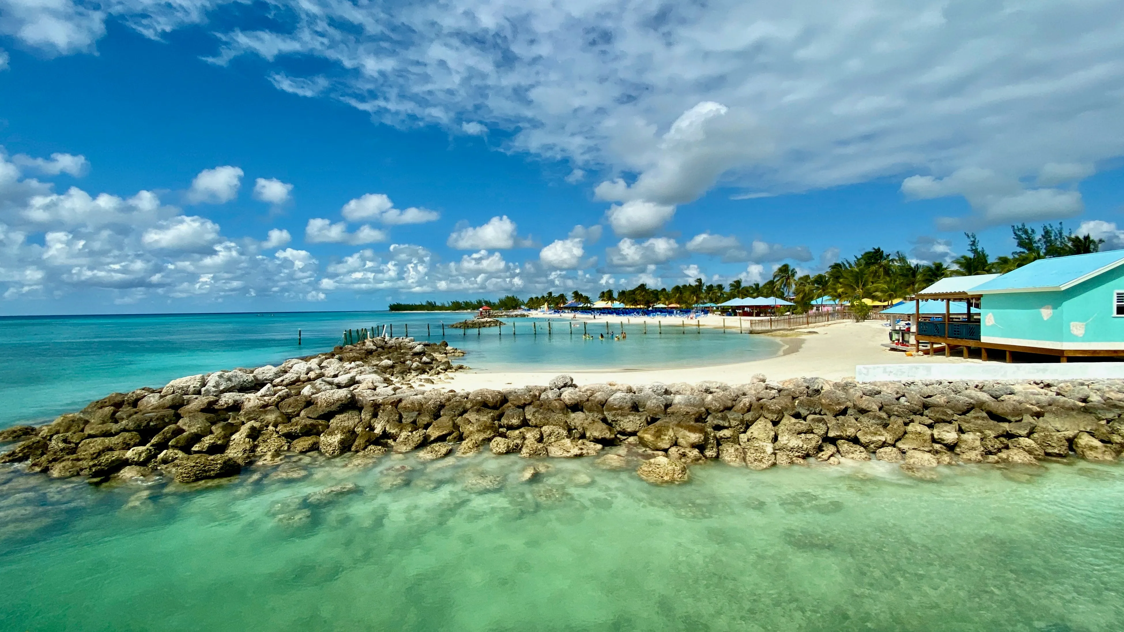 Turquoise water and a rock breakwater along a white sand beach lined with palms and cabanas on Eleuthera