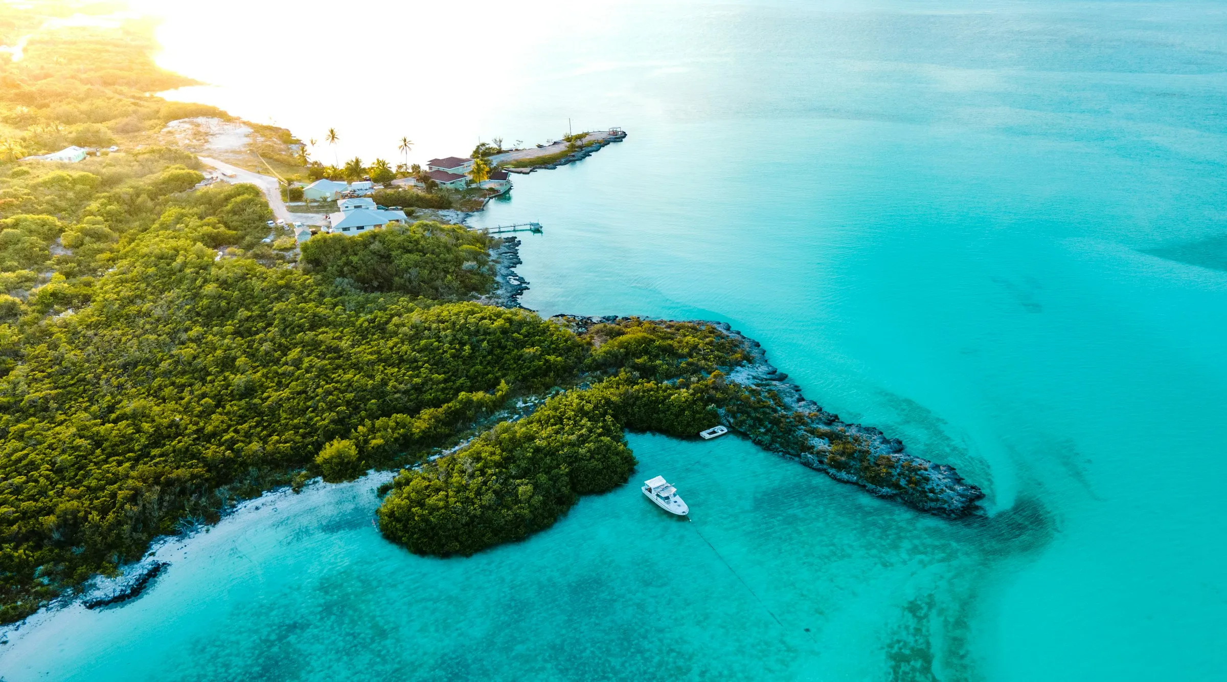 Aerial view of a green cay meeting turquoise shallows in the Exumas, with a small settlement, a dock, and a moored boat